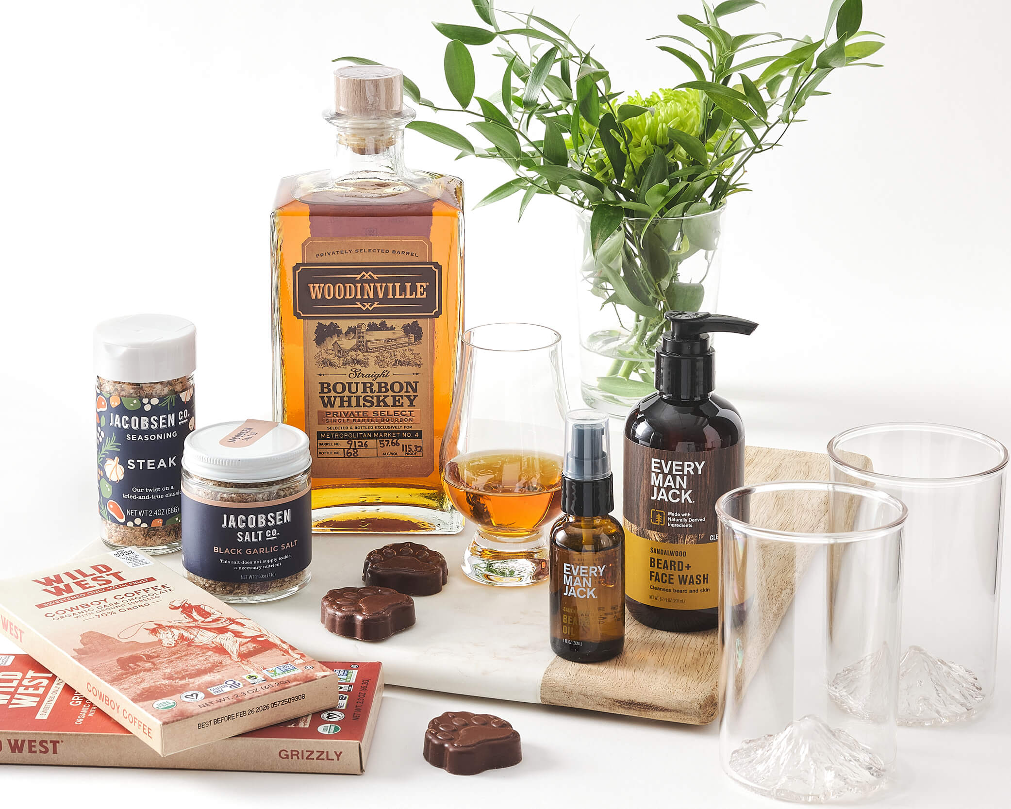 Whiskey, jarred seasonings, chocolate, facial care products, and greenery on a wooden cutting board