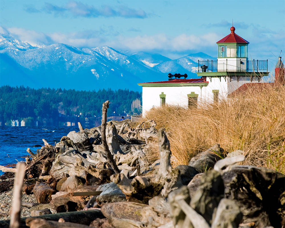 Image of a lighthouse on a rocky beach with snow capped mountains in the background.
