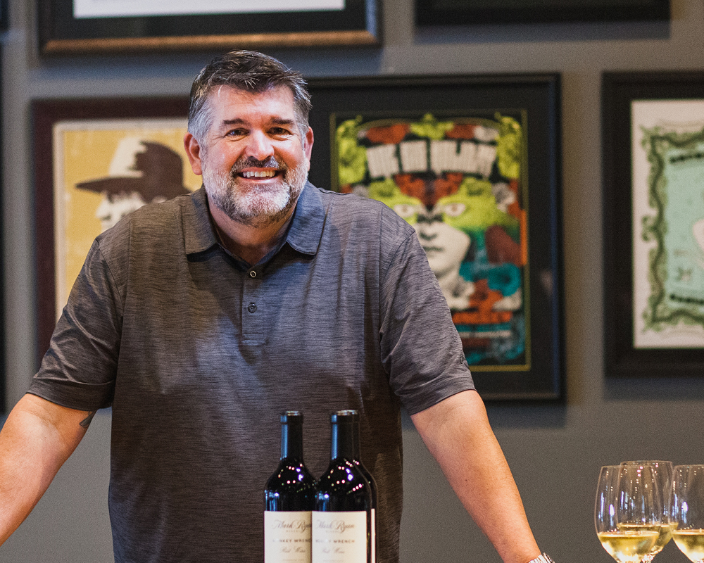 Mark Ryan smiling in wine tasting room with two bottles and two glasses on table