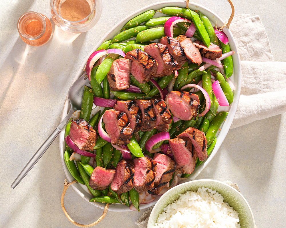 large platter of snap peas and red onions topped with sliced steak
