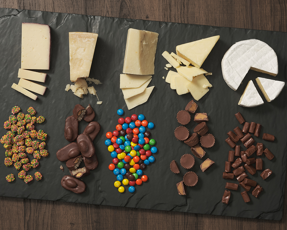 Five cheeses and five candies lined up on a slate board