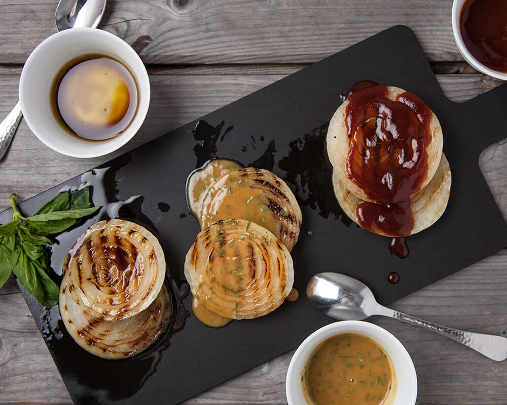 cutting board with 3 stacks of grilled onions topped with different sauces