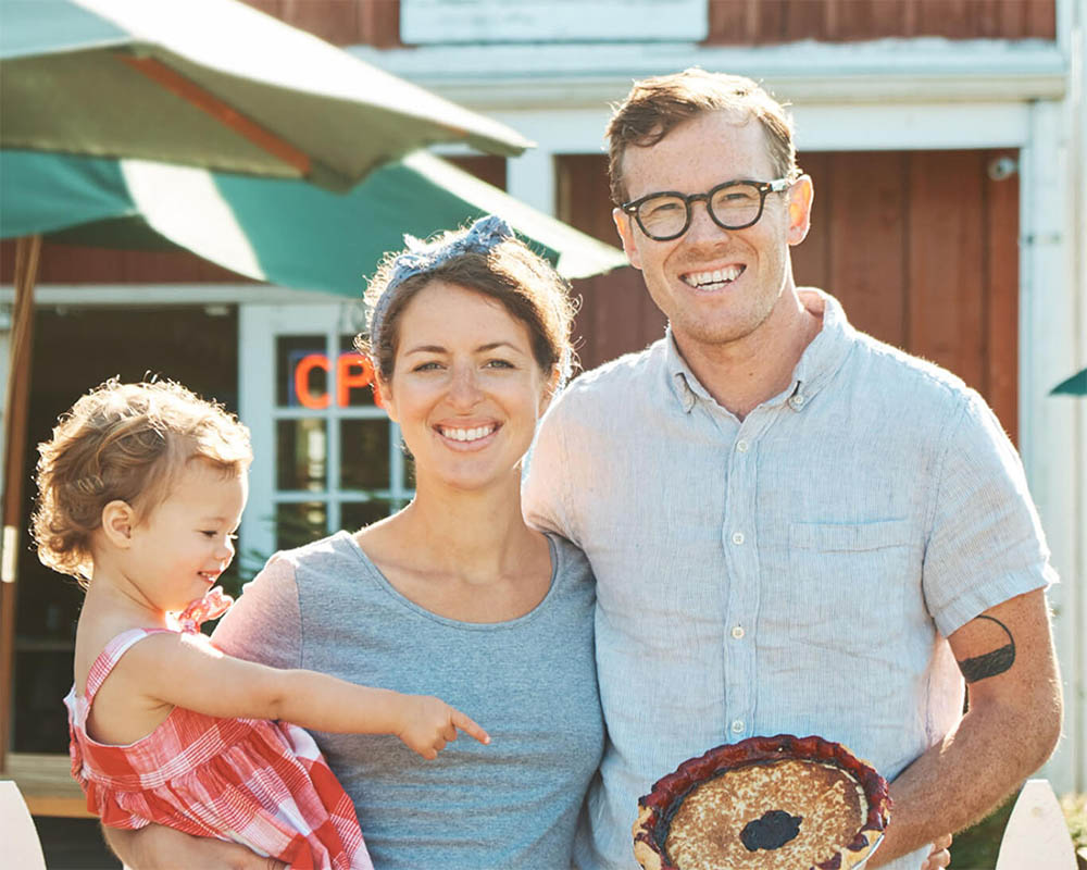 Joe Gunn holding a pie standing with his wife and child who is pointing to the pie