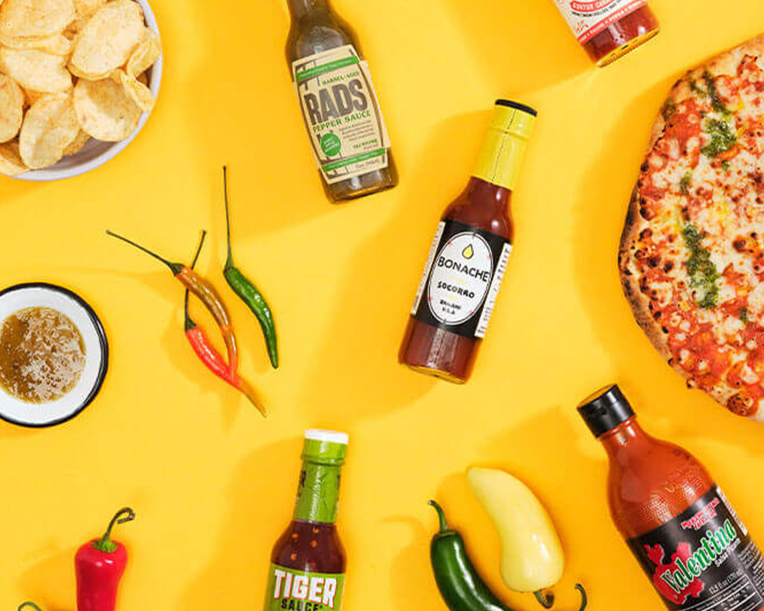 Five hot sauce bottles on a table with peppers chips and pizza