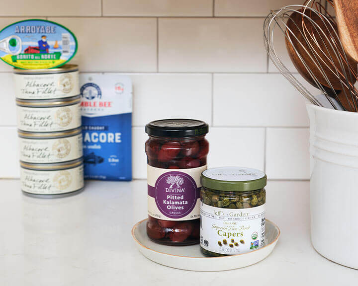 Jars of olives and capers on a pantry shelf.