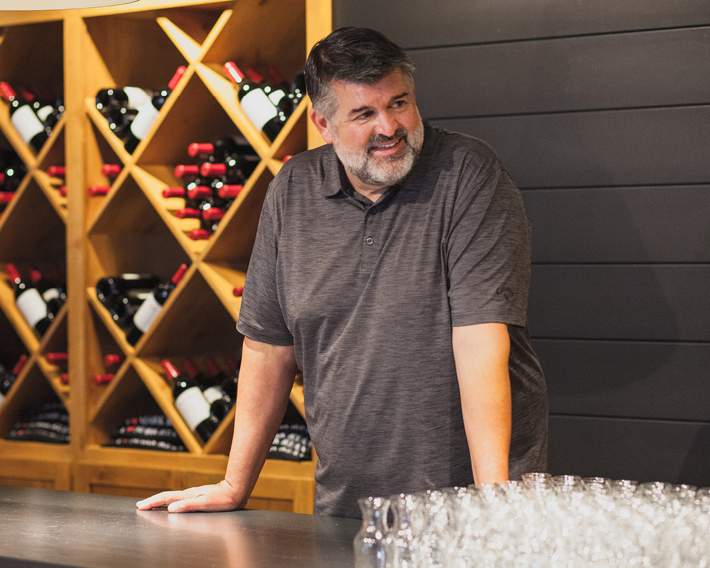 Mark Ryan leaning on table in wine tasting room with a wall of bottles behind him and rows of glasses in front of him.