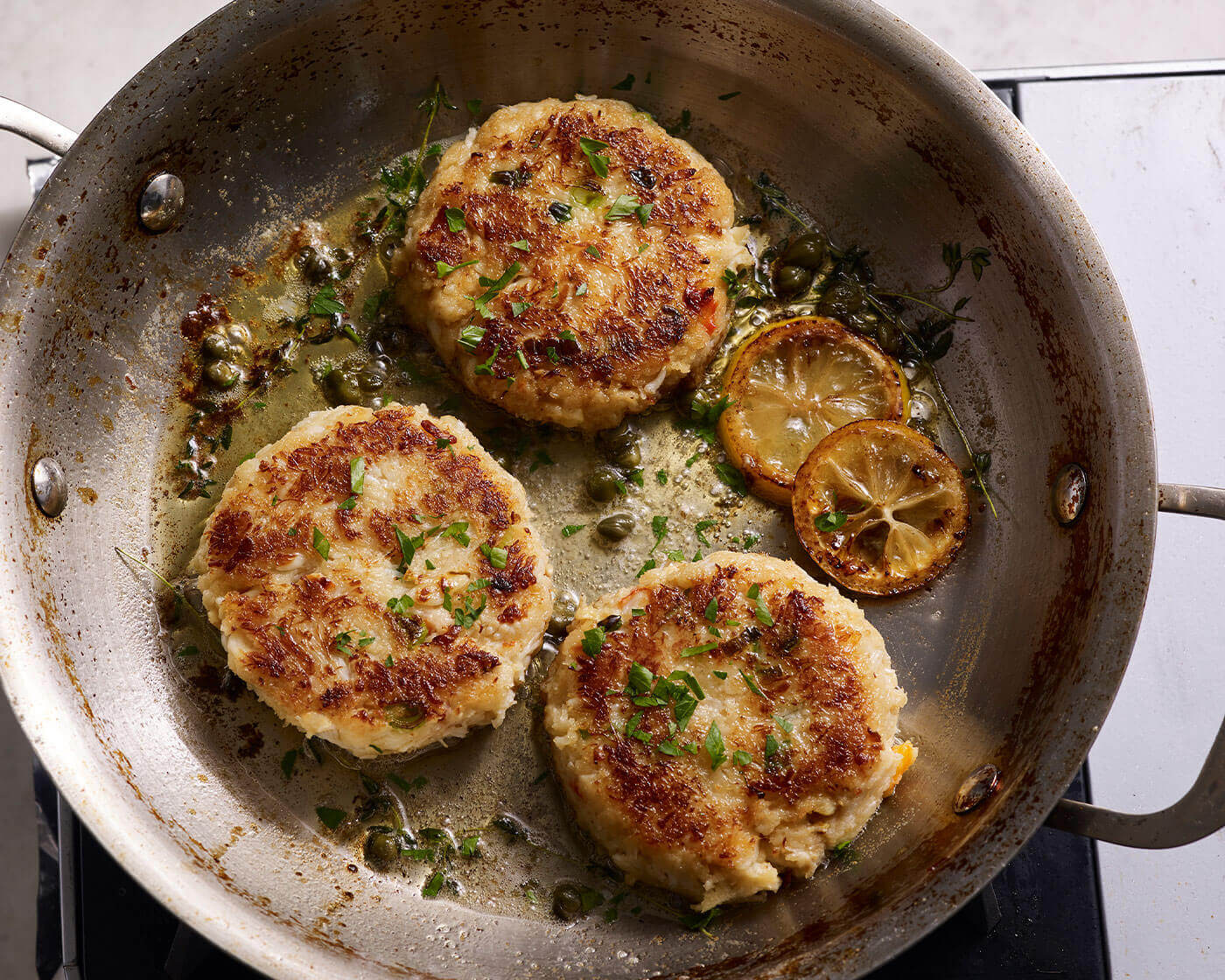 Crab cakes in a pan with lemon slices and capers