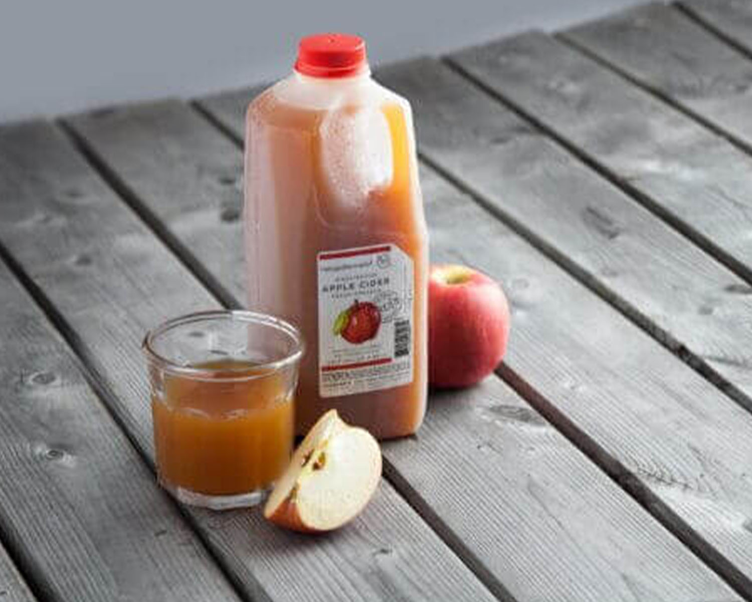 Jug of apple cider next to a glass of apple cider and a fresh apple on a wooden table.