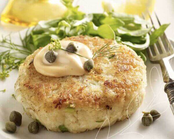 Crab cake topped with sauce and capers, served on a white plate with a salad.