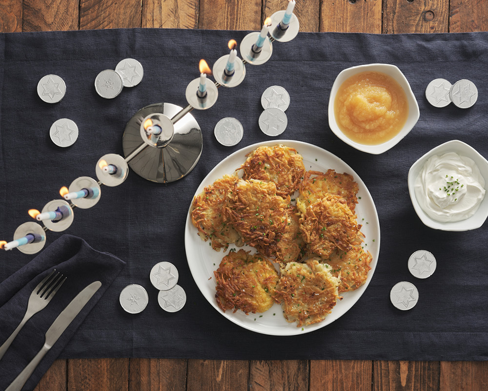 Latkes on a blue table runner with a applesauce, sour cream, a silver menorah, and gelt