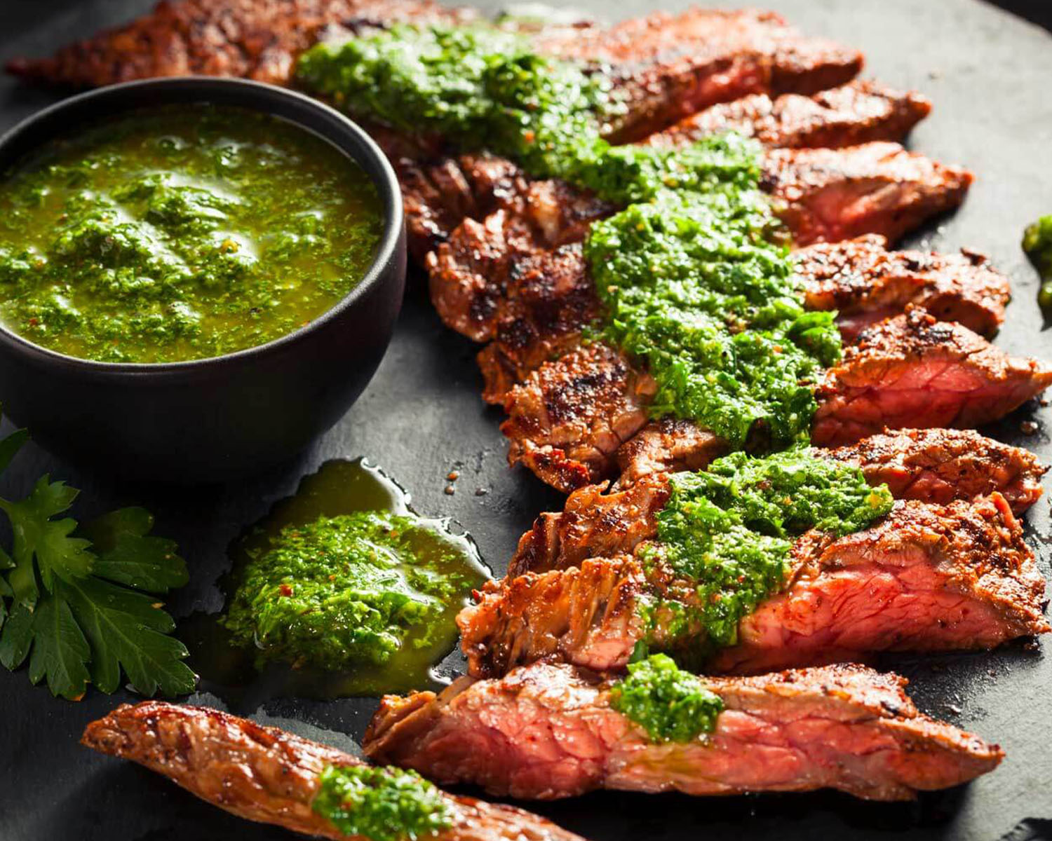 Green chimichurri sauce in a bowl and spread on top of a sliced steak.