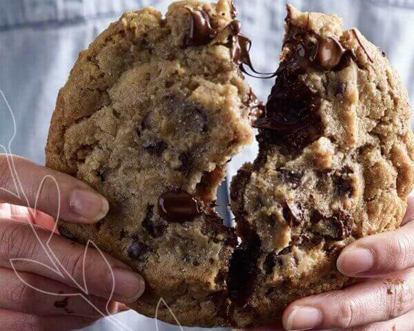 Hands breaking a giant cookie in half with melted chocolate chips.